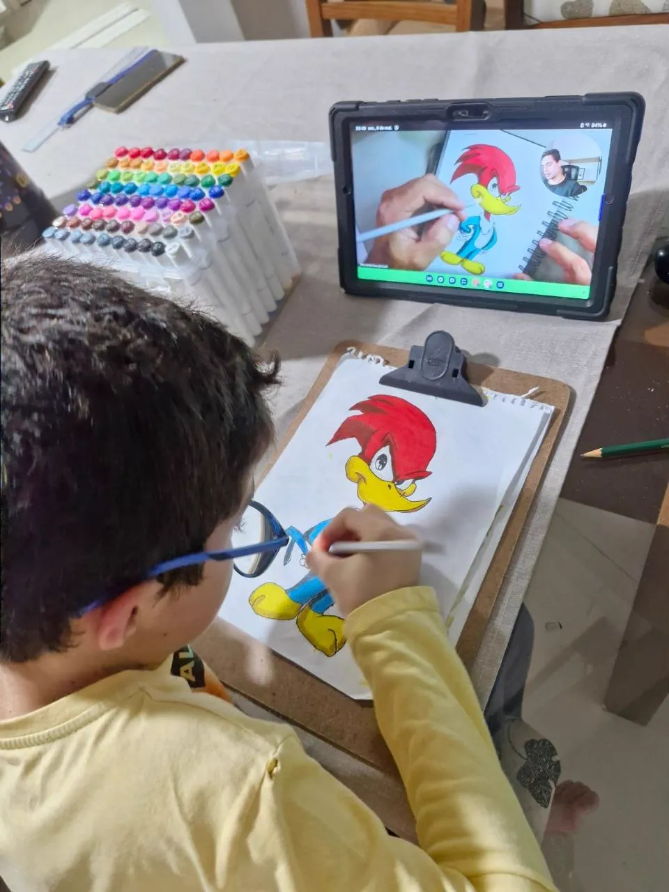 Menino desenhando o Pica-Pau no papel enquanto acompanha a aula ao vivo do Clube Line Art no tablet, com canetas coloridas na mesa