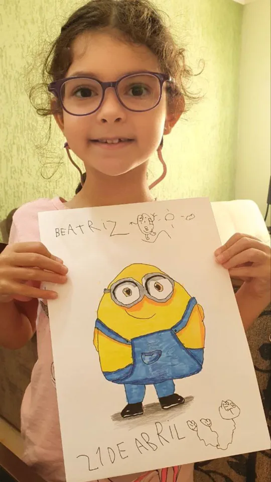 Menina de óculos sorrindo e mostrando o desenho do Minion que fez na aula ao vivo do Clube Line Art