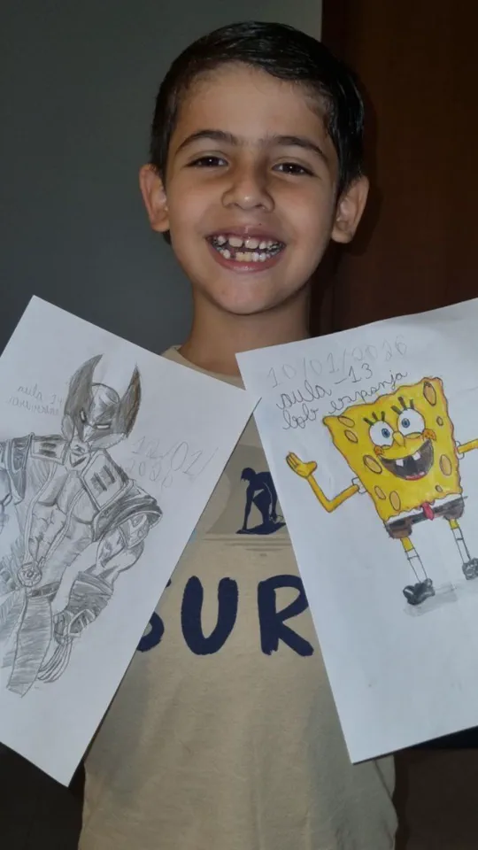 Menino sorrindo segurando dois desenhos que fez nas aulas: o Wolverine e o Bob Esponja